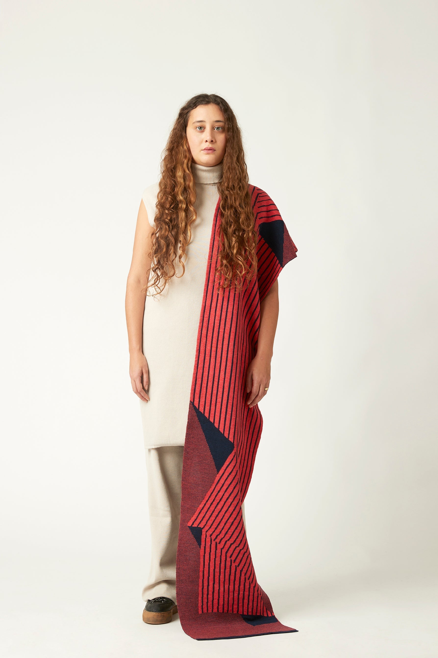 Wide shot of a young woman with a striped scarf draped over her shoulder. She's wearing a neutral light beige outfit and black shoes. The striped knitted scarf is coral pink and dark blue.